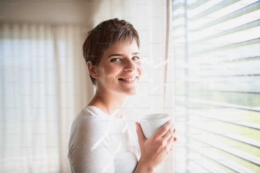 Retrato da jovem mulher feliz com café em pé pela janela dentro de casa em casa.