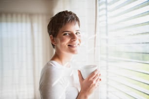 집에서 실내 창가에 커피를 들고 서 있는 젊은 행복한 여자의 초상화.