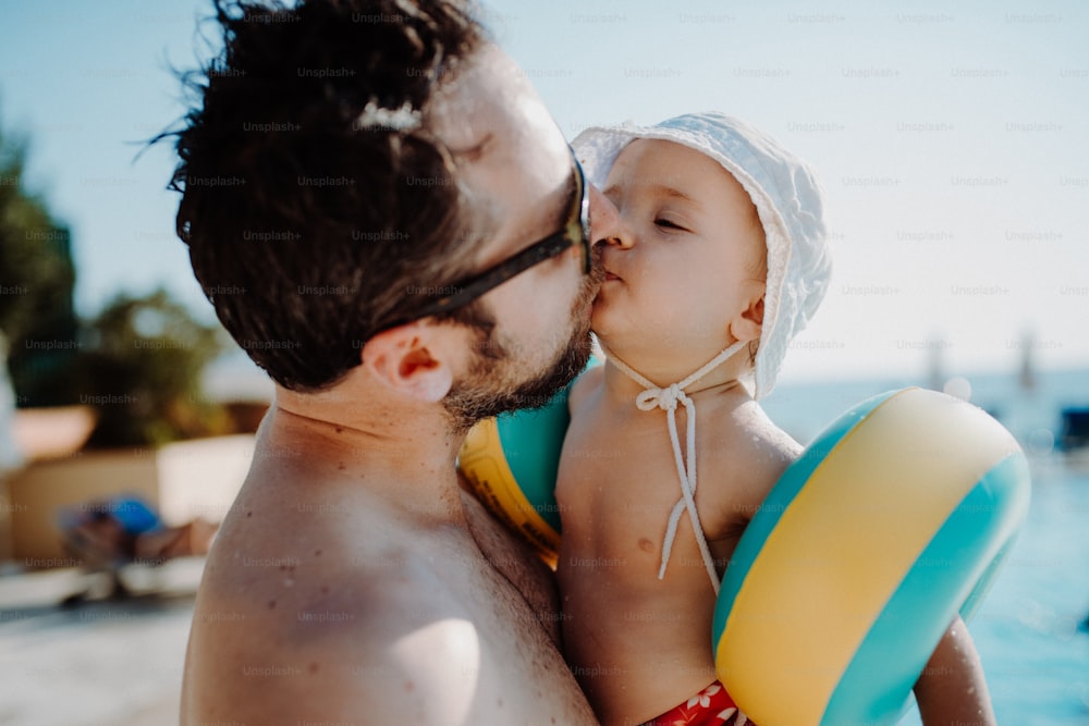 Un père avec un petit enfant avec des brassards debout au bord de la piscine pendant les vacances d’été.