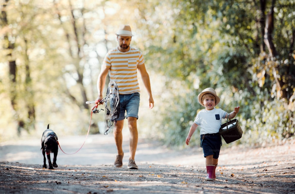 Um pai maduro com um filho pequeno e um cachorro andando em uma estrada na natureza, indo pescar.