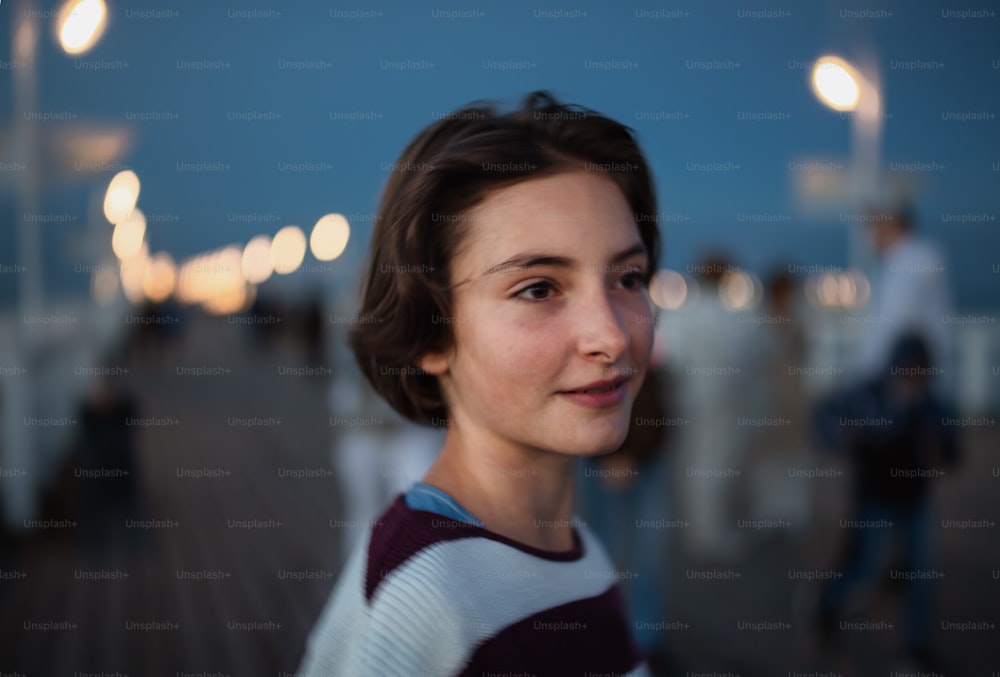 A portrait of preteen girl looking aside outdoors on pier by sea at dusk, holiday concept.
