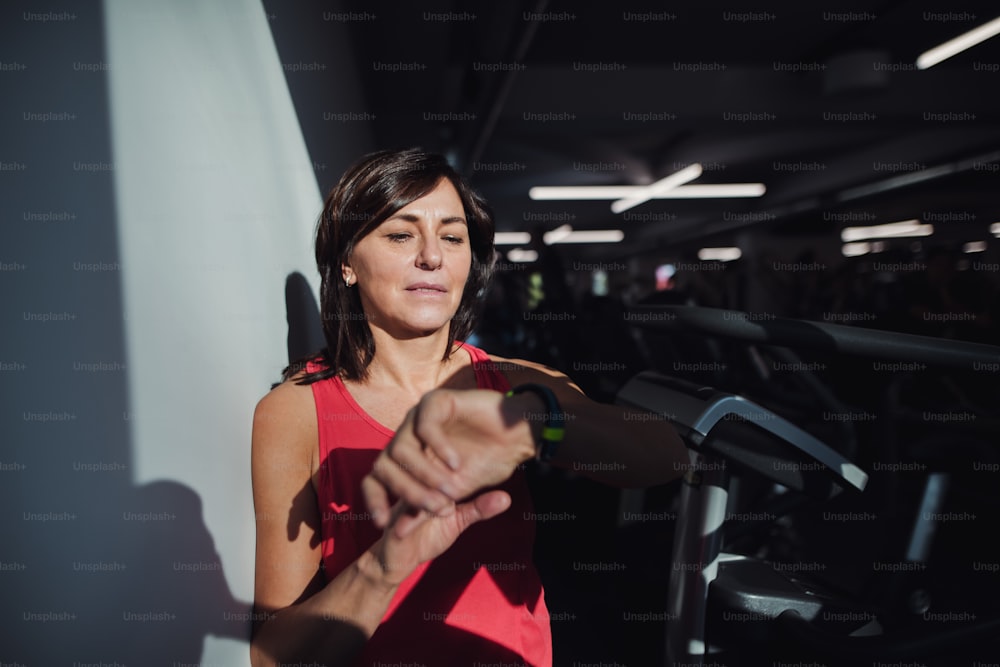 A content senior woman with smartwatch in gym resting after doing exercise, checking time. Copy space.