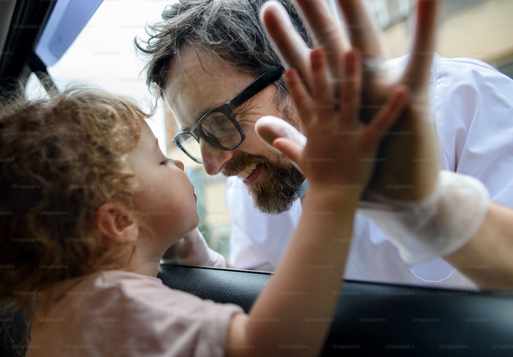 Doctor coming to see and greet small daugther in isolation, car window glass separating them.