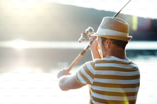 Un homme mûr avec un chapeau pêchant au bord d’un lac au coucher du soleil, tenant une canne à pêche. Espace de copie.
