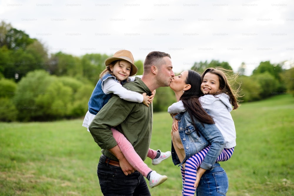 Familia feliz con dos hijas pequeñas de pie al aire libre en la naturaleza primaveral, besándose.