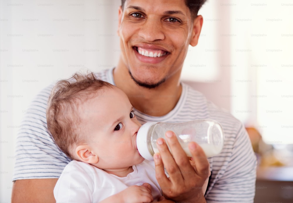 Un retrato de un padre alegre alimentando con biberón a un pequeño hijo pequeño en el interior de casa.