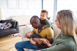 Beautiful young interracial family at home with their cute daughter and little baby son, sitting on couch.