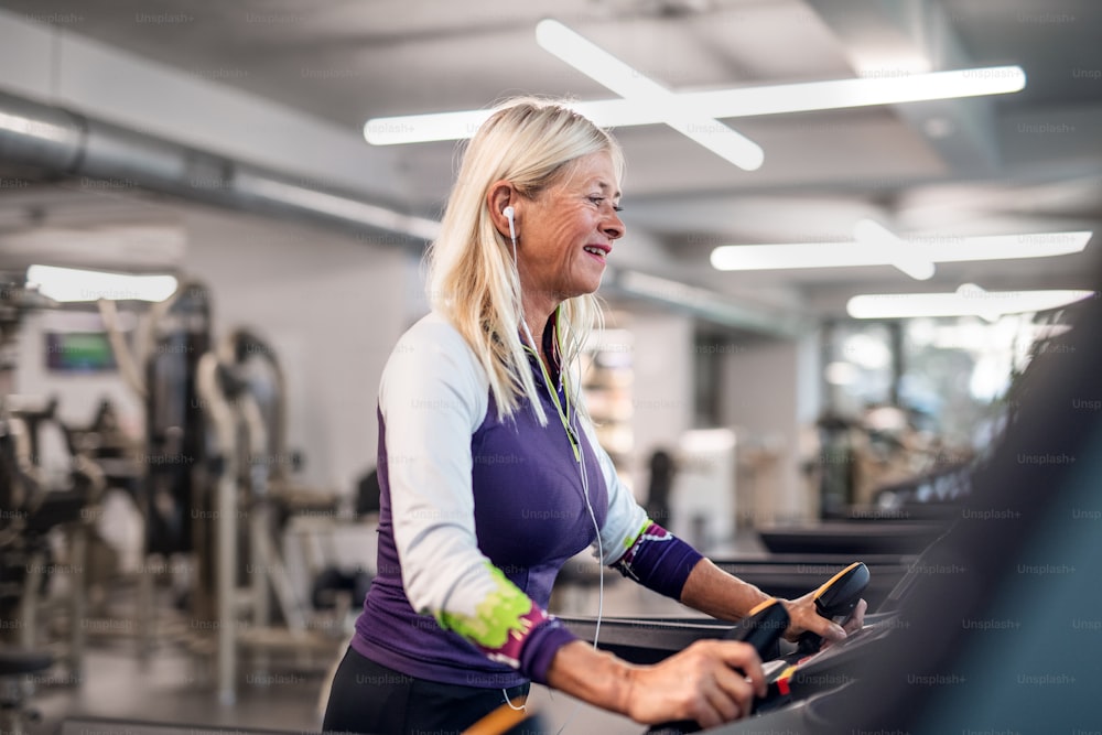 Eine glückliche ältere Frau mit Kopfhörern im Fitnessstudio, die Cardio-Training macht.