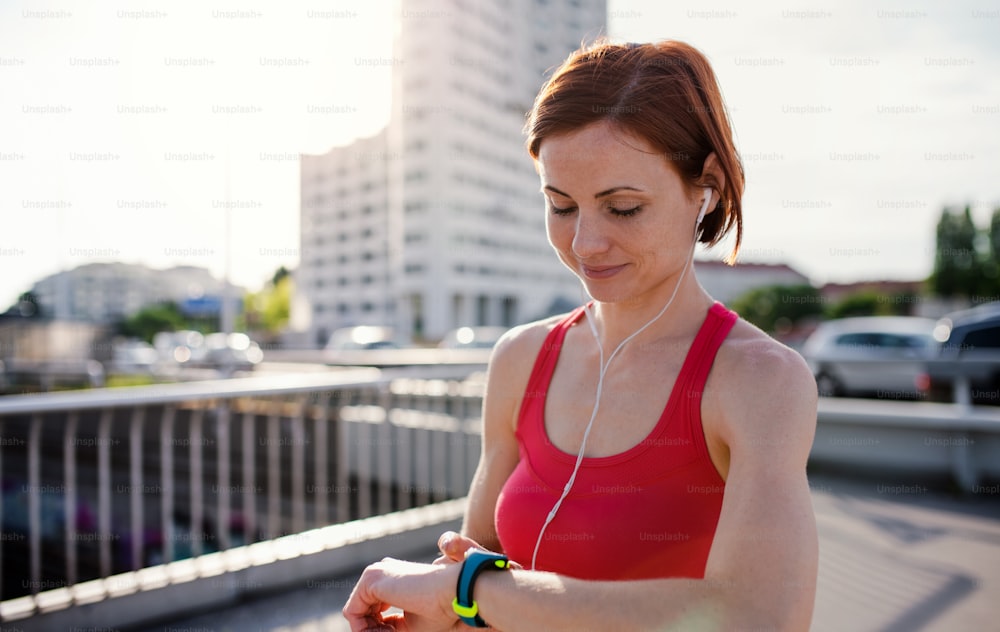 Giovane donna corridore con auricolari in città, usando lo smartwatch durante il riposo.