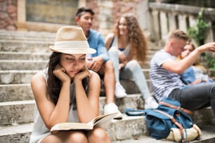 Attractive teenage student girl sitting on stone steps with her friends in front of university reading a book.