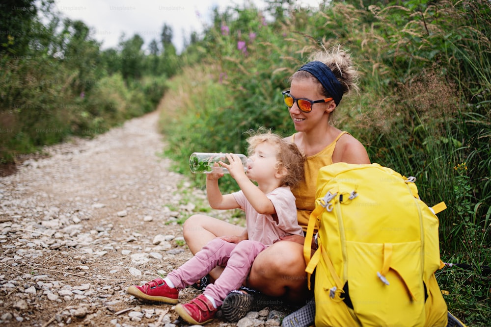 Mother with small toddler daughter hiking outdoors in summer nature, resting and drinking water.
