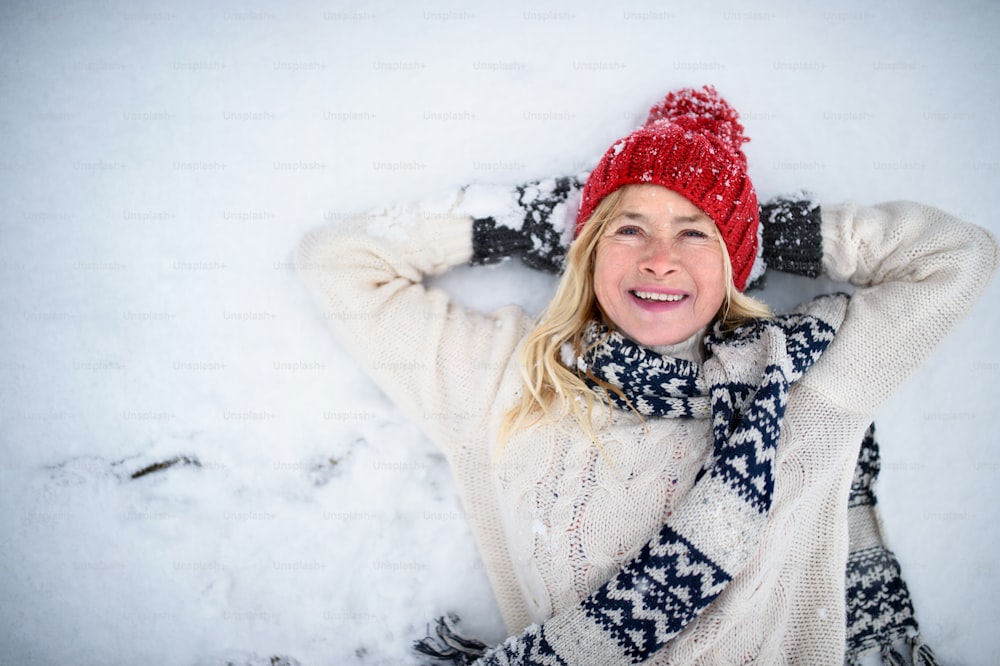 Top view portrait of happy senior woman with hat outdoors lying on snow, looking at camera.