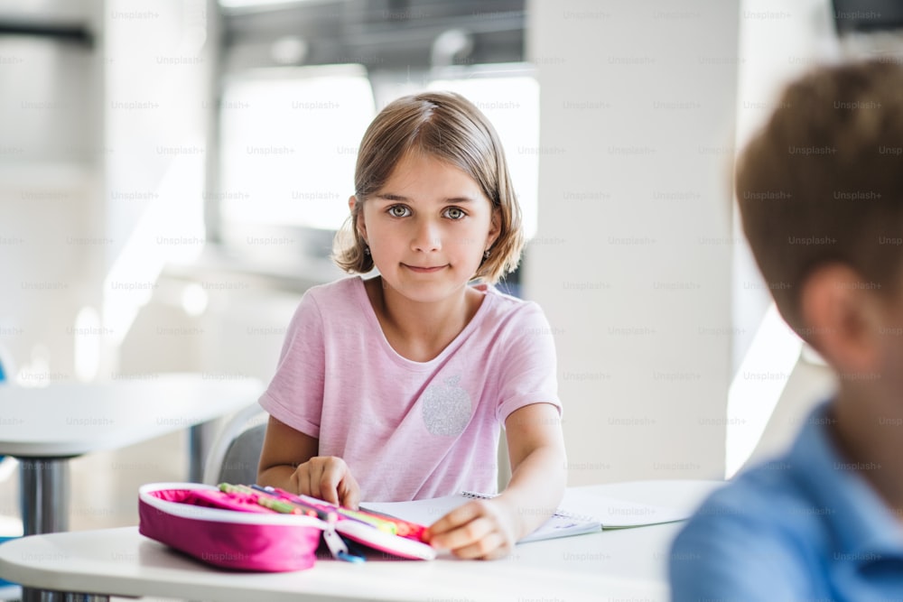Un retrato de una pequeña colegiala feliz sentada en el escritorio en el aula, mirando a la cámara.
