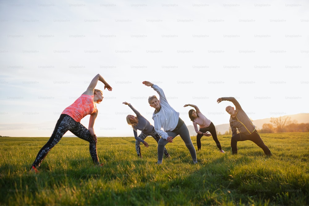 Un groupe de personnes âgées avec un instructeur de sport faisant de l’exercice à l’extérieur dans la nature au coucher du soleil, un mode de vie actif.