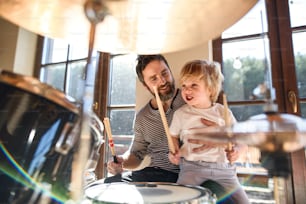 Happy small boy with father indoors at home, playing drums.