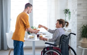 Portrait of disabled mature woman in wheelchair sitting at the table with a son indoors at home, eating.