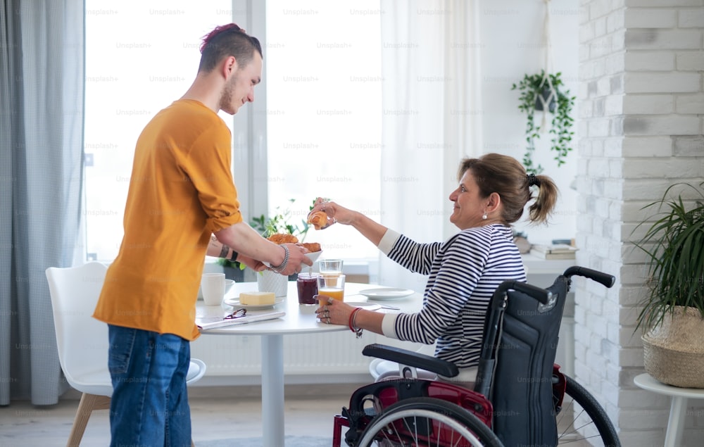 Portrait of disabled mature woman in wheelchair sitting at the table with a son indoors at home, eating.