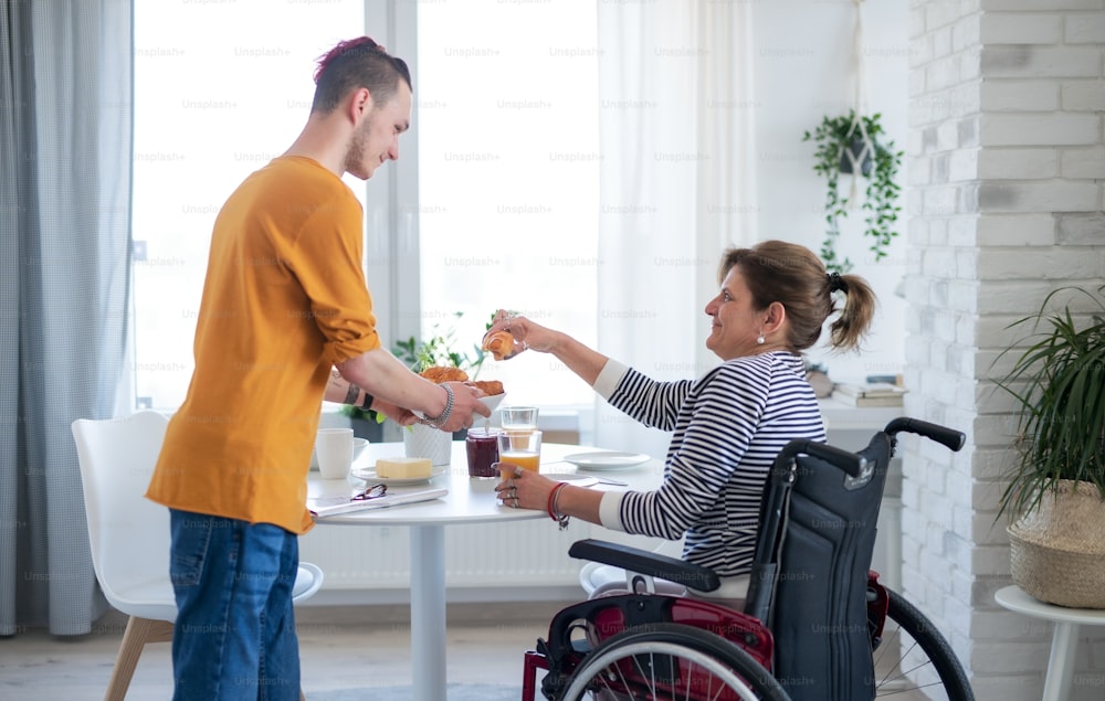 Portrait of disabled mature woman in wheelchair sitting at the table with a son indoors at home, eating.