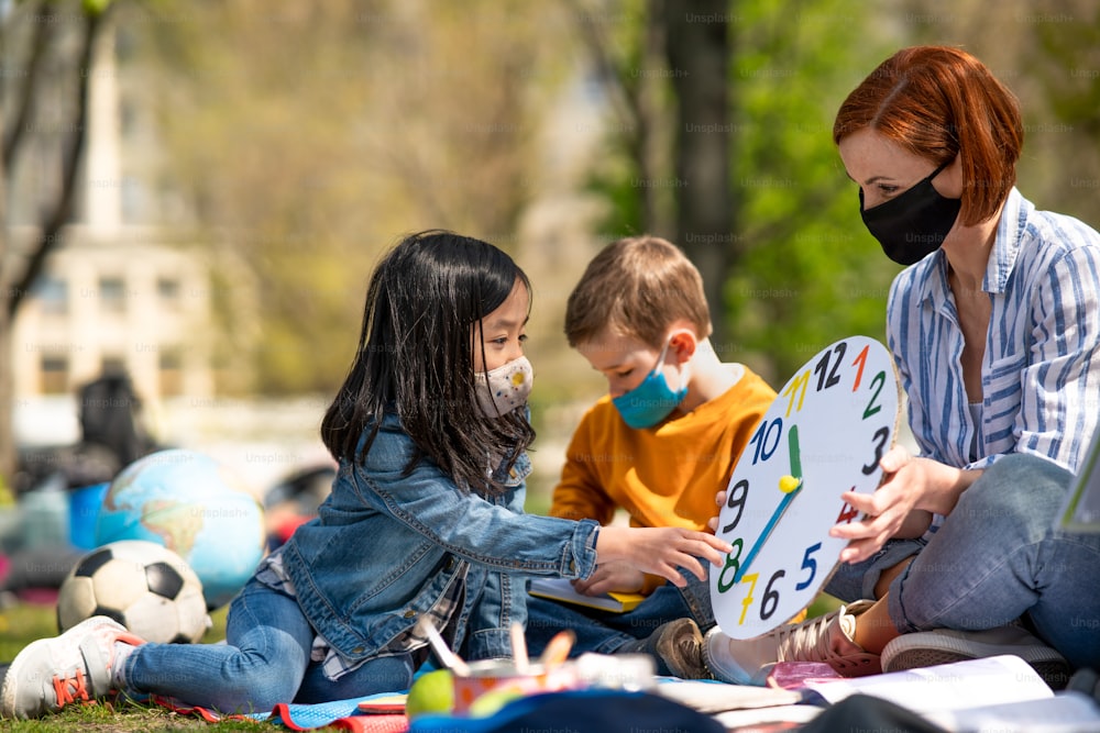 Ein Lehrer mit kleinen Kindern, die draußen im Stadtpark sitzen, Lerngruppenunterricht und Coronavirus-Konzept.