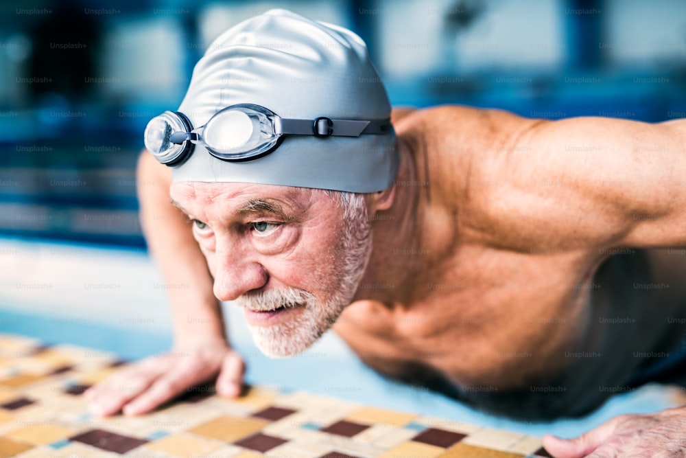 Homme âgé debout près de la piscine intérieure, s’étirant. Retraité actif qui fait du sport.