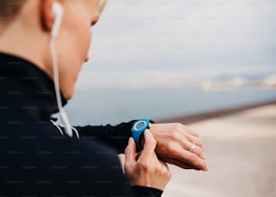 A midsection of young sportswoman with earphones standing outdoors on beach, using smartwatch.