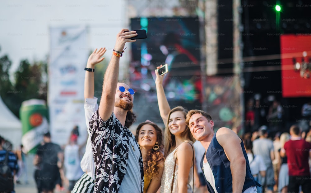 Vista frontale del gruppo di giovani amici con smartphone al festival estivo, scattando selfie.