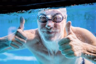 屋内プールで水中を泳ぐ年配の男性。スポーツを楽しむ現役の年金生活者。親指を立てるジェスチャー。