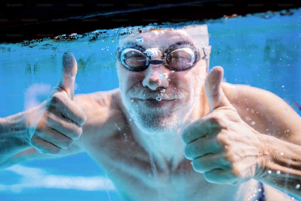 Homme âgé nageant sous l’eau dans une piscine intérieure. Retraité actif qui fait du sport. Geste du pouce levé.