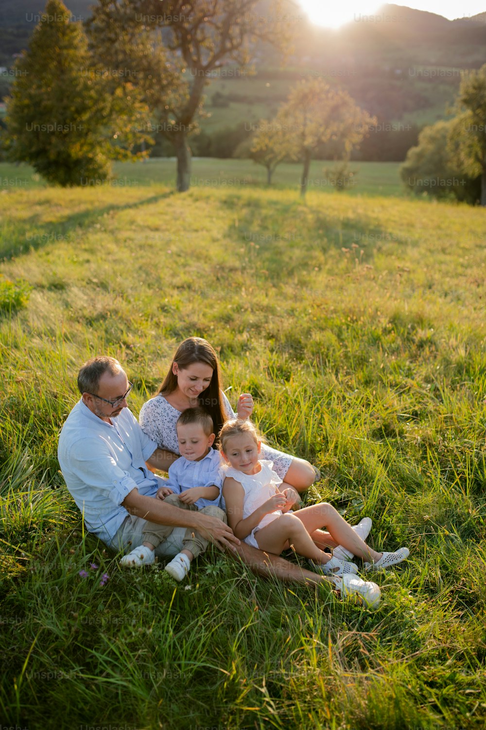 Familia joven feliz con dos niños pequeños sentados en el prado al aire libre al atardecer.