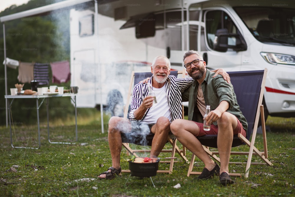 A mature man with senior father looking at camera at campsite outdoors, barbecue on caravan holiday trip.