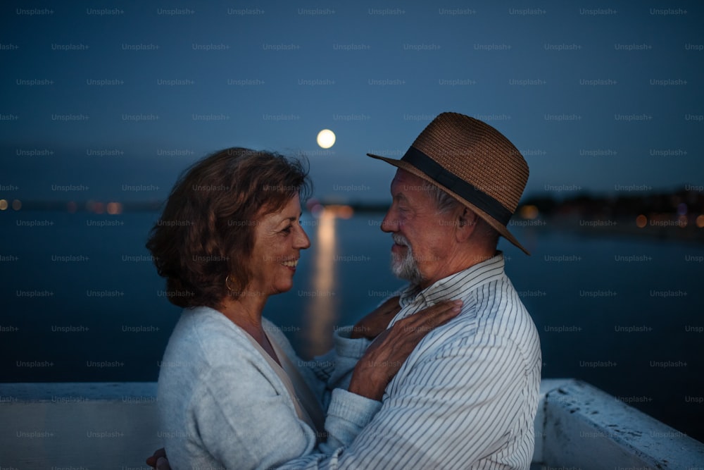 Una feliz pareja de ancianos abrazados al aire libre en el muelle junto al mar al anochecer, mirándose.