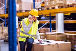 Young female warehouse worker with smartphone. A woman or a supervisor sitting on boxes, making a phone call.
