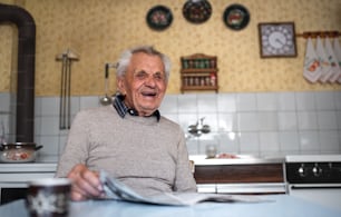 A portrait of elderly man sitting at the table indoors at home, reading newspapers.