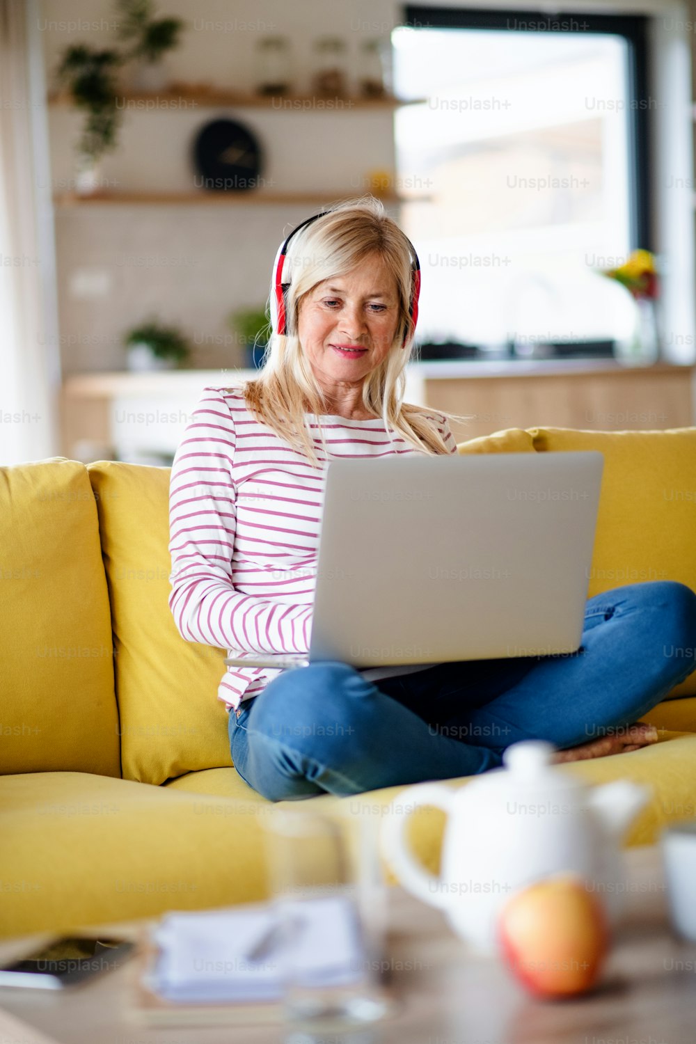 Femme âgée avec des écouteurs et un ordinateur portable assise sur un canapé à l’intérieur à la maison, se détendant.