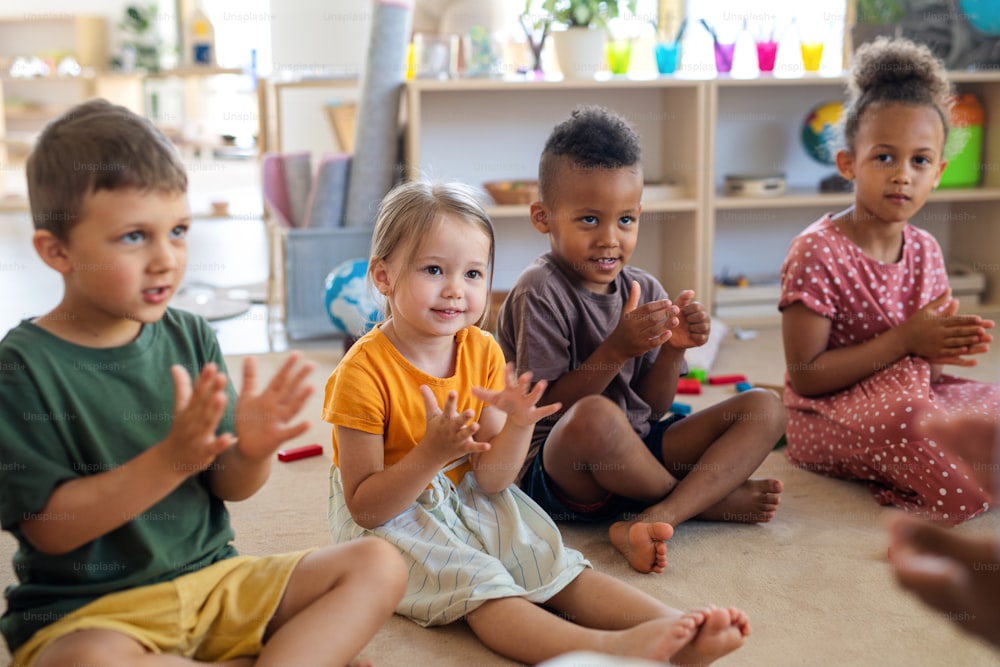 Um grupo de pequenas crianças da creche sentadas no chão dentro de casa na sala de aula, batendo palmas.