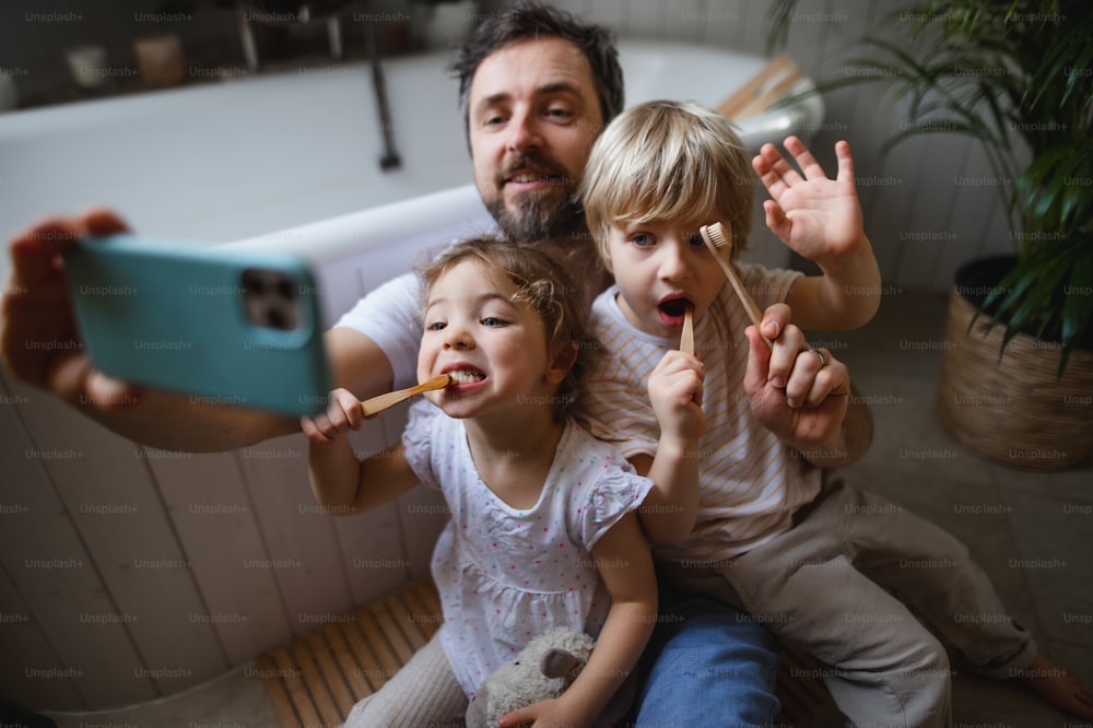 Mature father with two small children brushing teeth indoors at home, taking selfie.