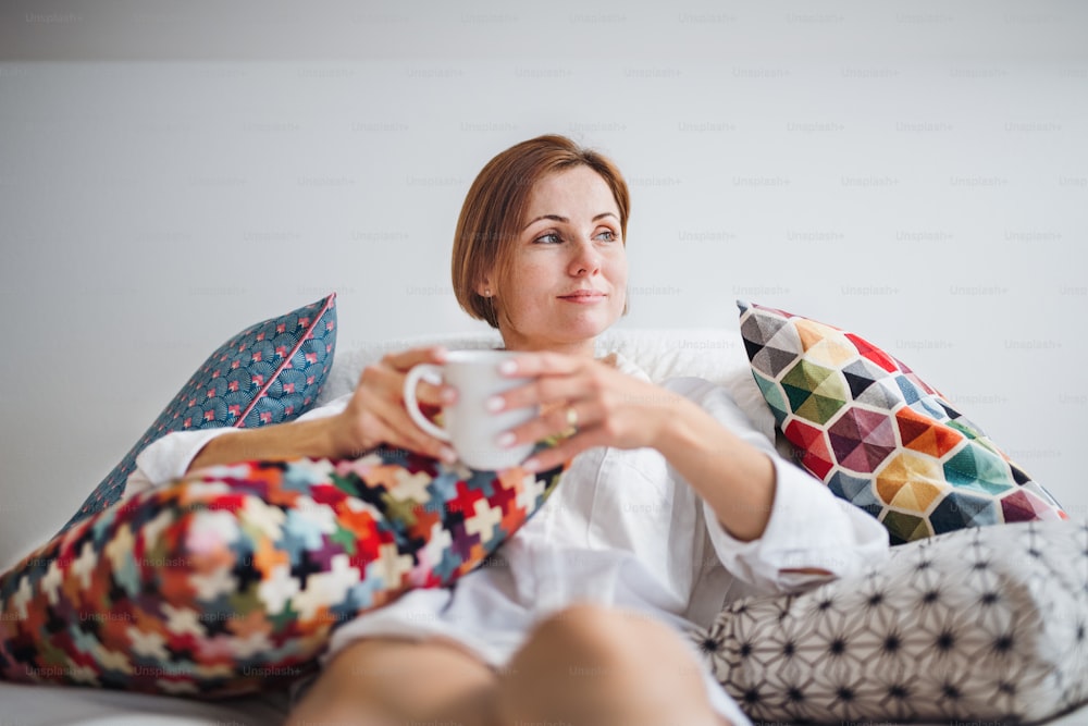 Young woman with night shirt sitting indoors on bed in the morning, holding cup of coffee.