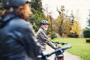 An active senior couple with helmets and electrobikes standing outdoors on a road in nature.