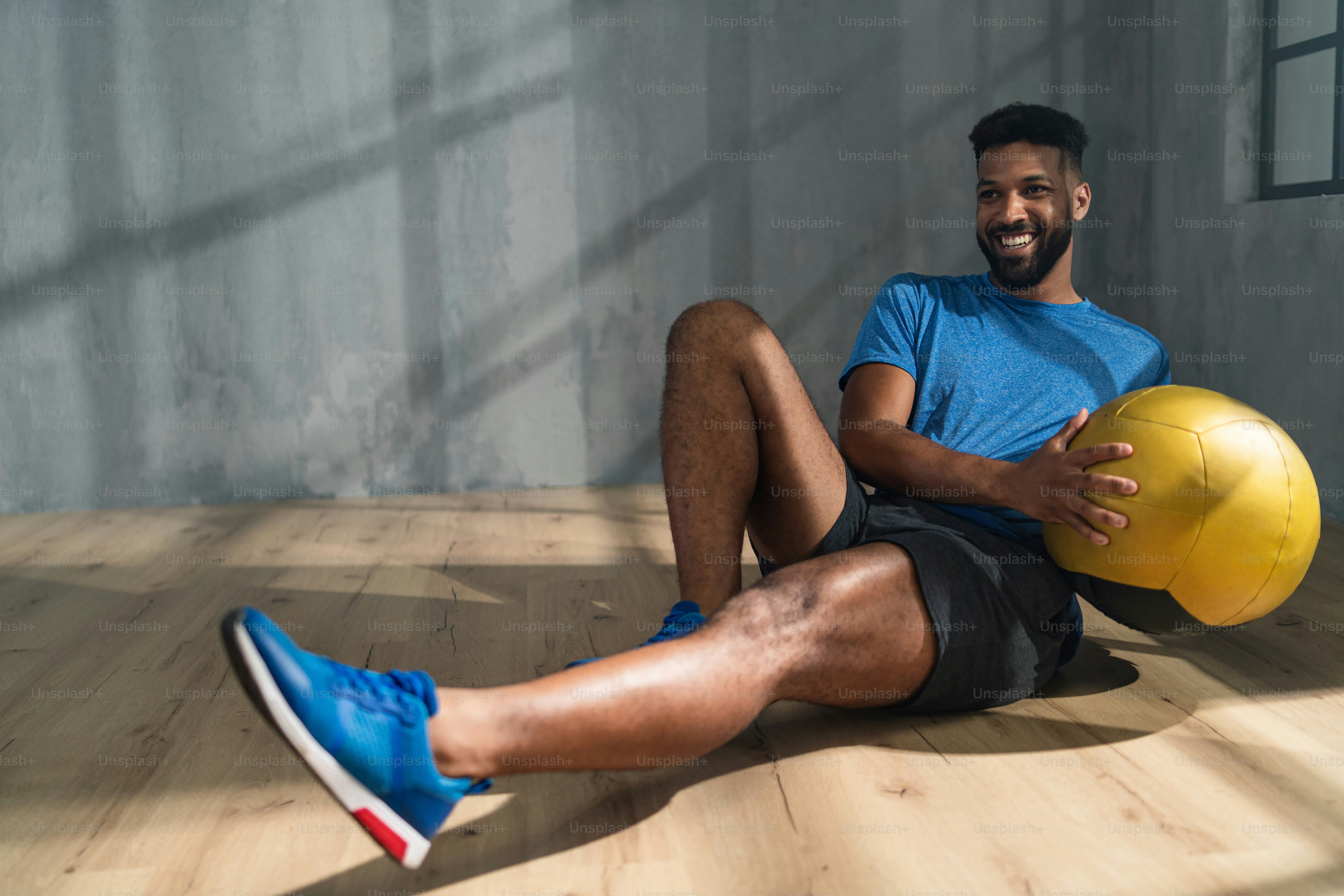 A young African American sportsman sitting and lifting a medicine ball indoors, workout training concept.