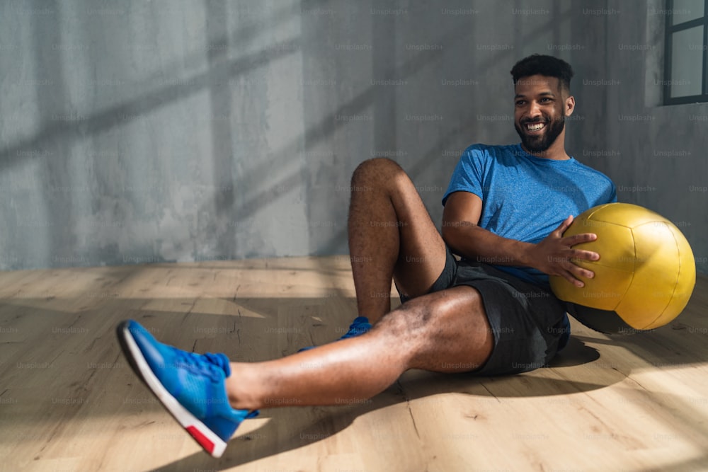 Un jeune sportif afro-américain assis et soulevant un ballon médicinal à l’intérieur, concept d’entraînement d’entraînement.