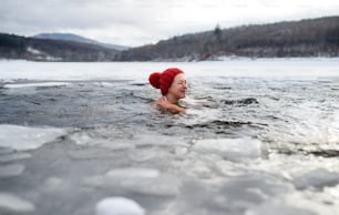 A happy active senior woman in hat diving outdoors in water in winter, cold therapy concept. Copy space.