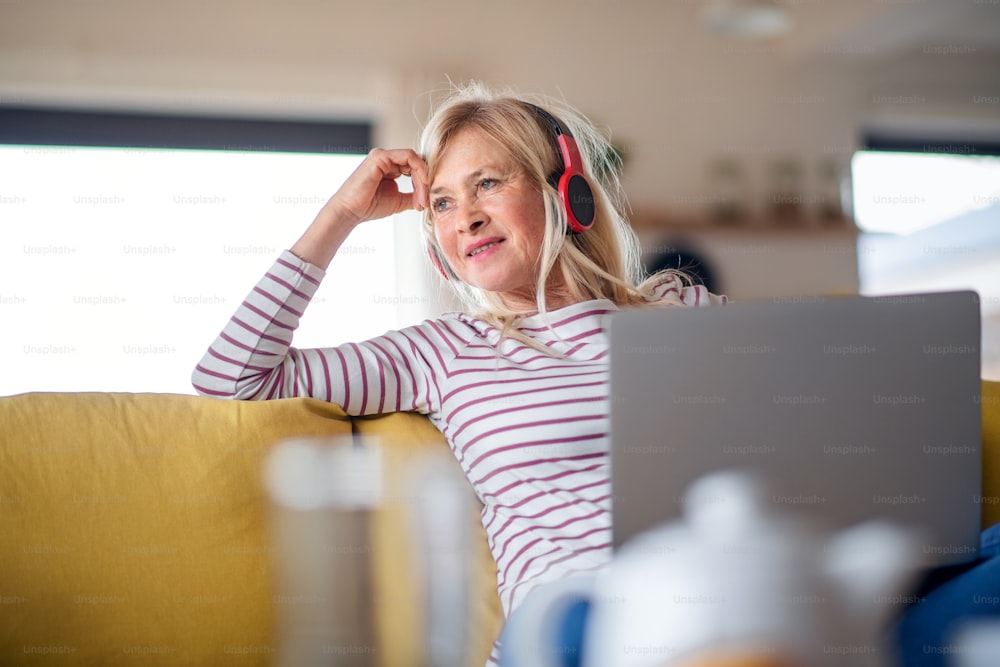Senior woman with headphones and laptop sitting on sofa indoors at home, relaxing.