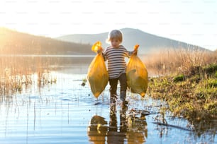 Niño pequeño recogiendo basura al aire libre junto al lago en la naturaleza, concepto de plogging.