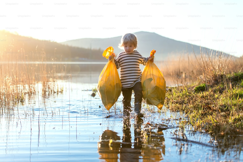 Bambino piccolo che raccoglie la spazzatura all'aperto dal lago nella natura, concetto di plogging.