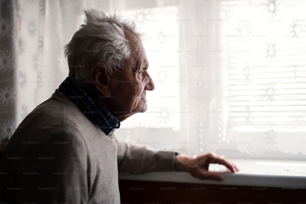 A portrait of elderly man standing indoors at home, looking out through window.