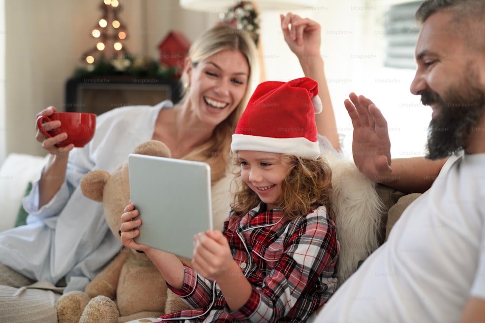 Famille avec petite fille à l’intérieur à la maison à Noël, ayant un appel vidéo sur tablette.