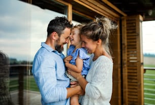 Happy family with small daughter standing on patio of wooden cabin, holiday in nature concept.
