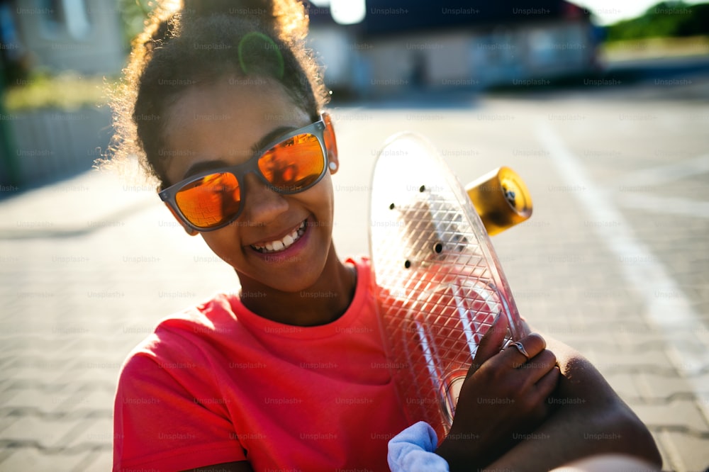 Front view of teenager girl with sunglasses and skateboard outdoors in city, looking at camera.