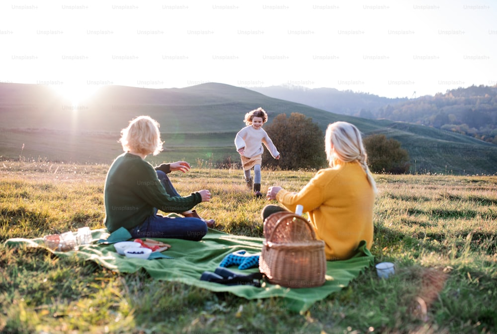 Niña pequeña feliz con madre y abuela haciendo un picnic en la naturaleza al atardecer.