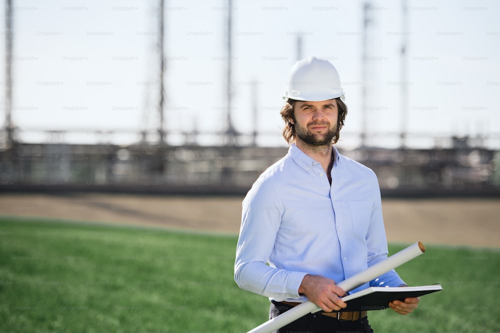 Young engineer with hard hat standing outdoors by oil refinery, looking at camera.